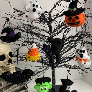 May include: A black twig tree decorated with felt Halloween ornaments. The ornaments include a ghost, a witch's hat, a pumpkin, a candy corn, a black cat, a bat, a Frankenstein, a witch's boot, and a ghost with a banner.