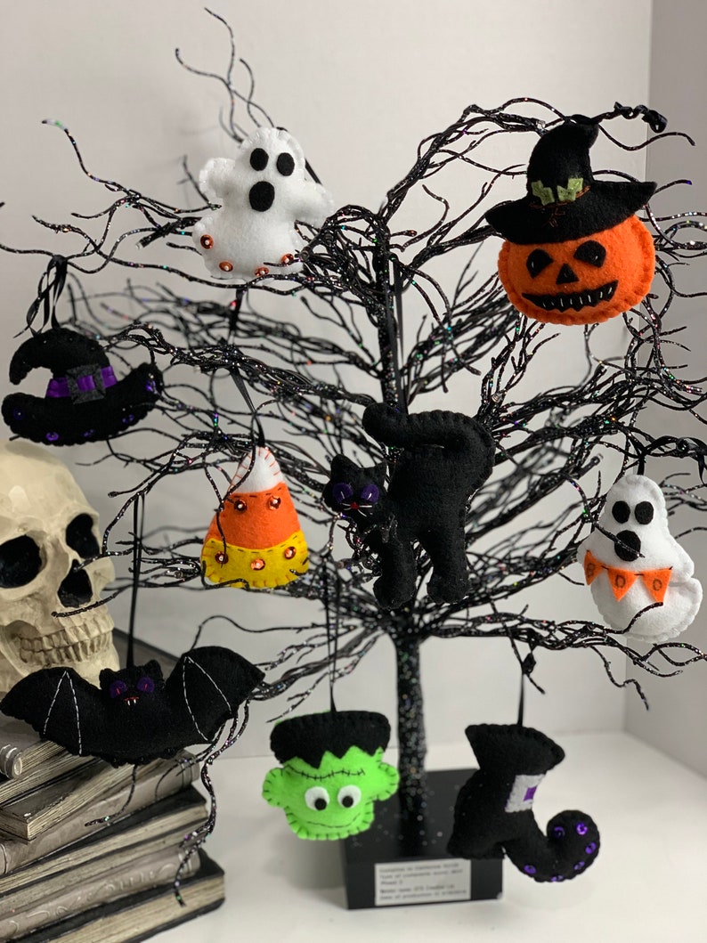 May include: A black twig tree decorated with felt Halloween ornaments. The ornaments include a ghost, a witch's hat, a pumpkin, a candy corn, a black cat, a bat, a Frankenstein, a witch's boot, and a ghost with a banner.