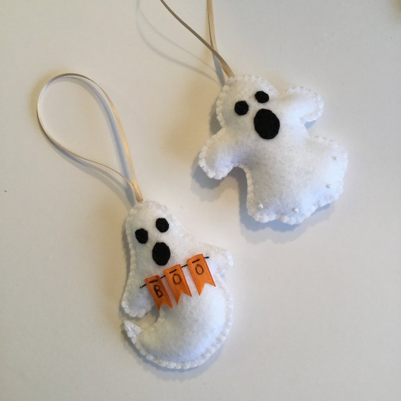 May include: Two white felt ghost ornaments with black eyes and mouths. One ghost has a small orange banner that says "BOO" hanging from it.