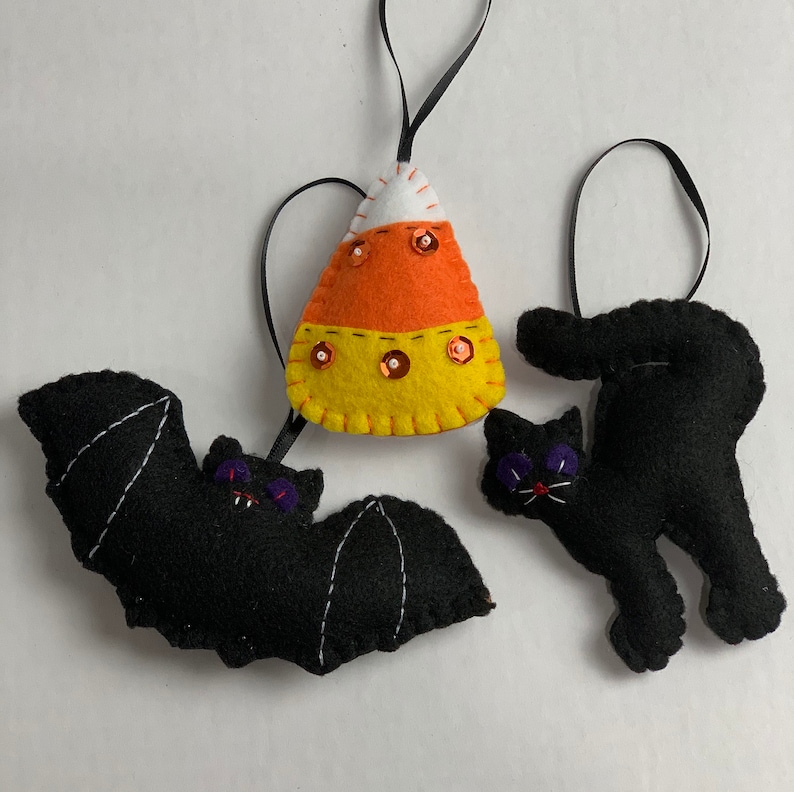 May include: Three felt Halloween ornaments: a black bat with purple eyes, a black cat with purple eyes, and a candy corn with orange, yellow, and white stripes and gold sequins.