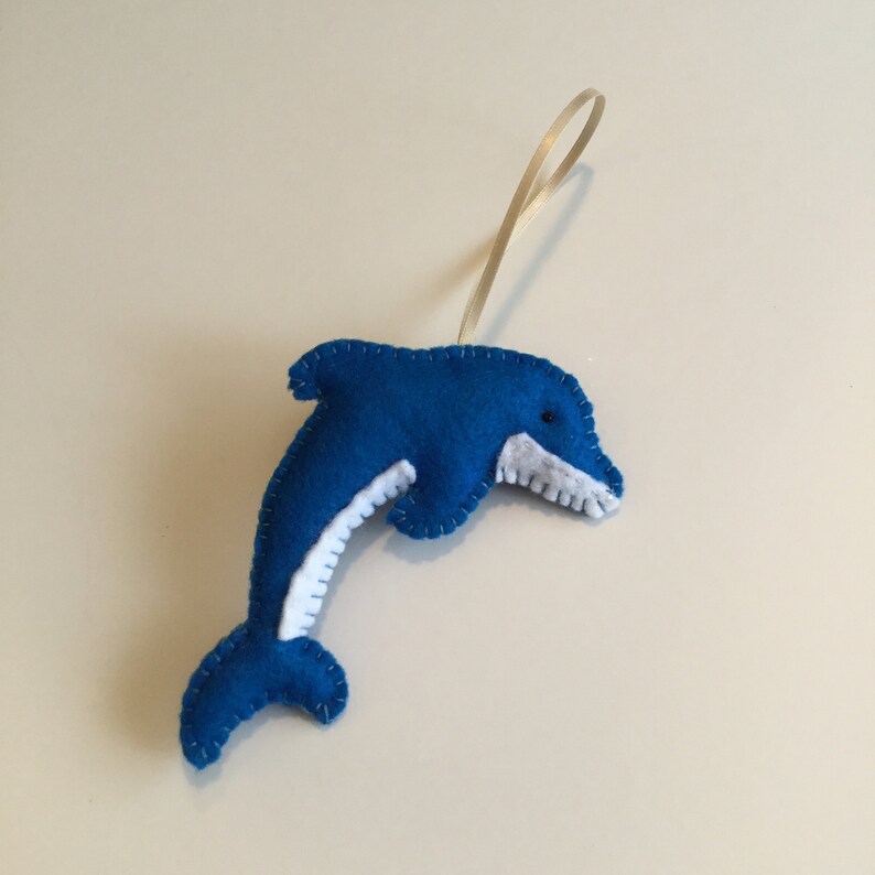 May include: A blue felt dolphin ornament with white felt accents and black eyes. The dolphin is hanging from a cream-colored ribbon.