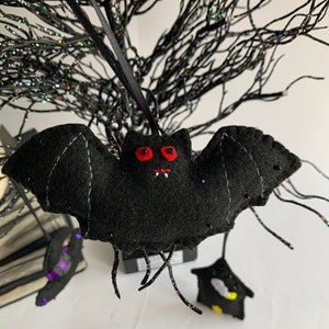 May include: A black felt bat with red eyes and white teeth hanging from a black string. The bat is hanging from a black twig-like branch.