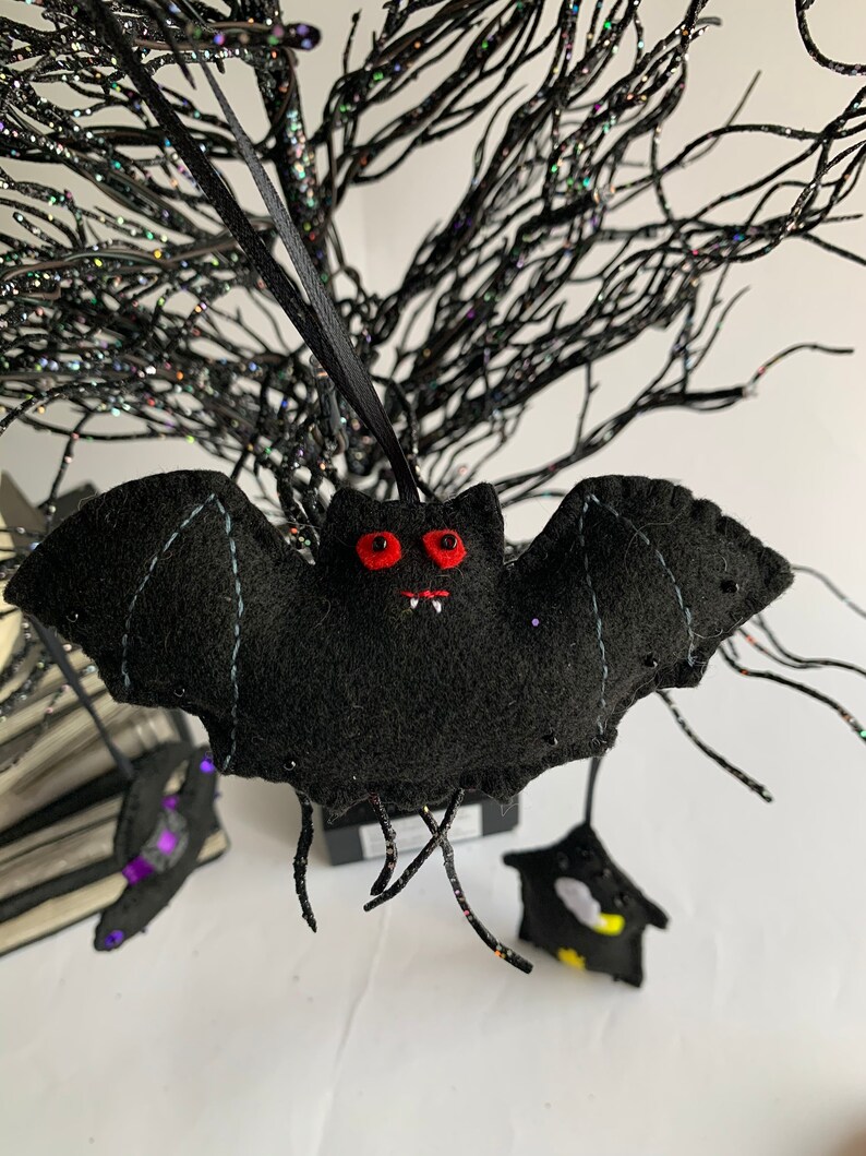 May include: A black felt bat with red eyes and white teeth hanging from a black string. The bat is hanging from a black twig-like branch.