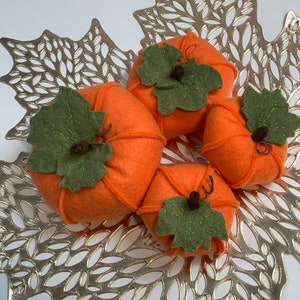 May include: Four felt pumpkins in orange with green felt leaves and stems. The pumpkins are arranged on a gold leaf-shaped placemat.
