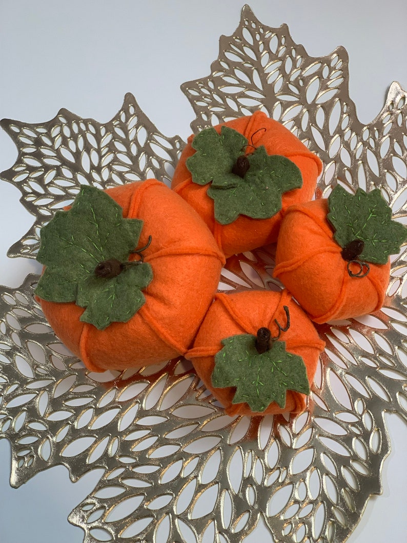 May include: Four felt pumpkins in orange with green felt leaves and stems. The pumpkins are arranged on a gold leaf-shaped placemat.
