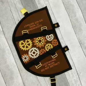 May include: Brown, semi-circular organizer with black trim and gold hardware. The organizer features a design of gold, silver, and bronze gears. The text "CLOCKWORKS AT WORK DO NOT DISTRACT" is embroidered on the brown fabric.