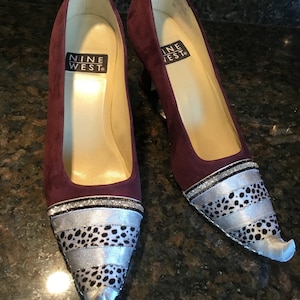 May include: A pair of burgundy suede high heels with a pointed toe. The shoes have a silver and black patterned trim around the toe and a silver glitter band.