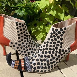 May include: A pair of black and white polka dot heeled boots with orange suede accents. The boots have a pointed toe and a witch's shoe design with a pointed toe and a curved, pointed heel.