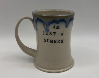 May include: A white ceramic mug with a blue and brown glaze. The mug has a handle and says "I AM JUST A NUMBER" in black lettering.