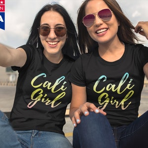K&ouml;nnte beinhalten: Zwei Personen tragen schwarze T-Shirts mit dem Text "Cali Girl" in einem Regenbogen-Verlauf. Der Text ist in einer handschriftlichen Schriftart. Die Personen l&auml;cheln und tragen Sonnenbrillen.