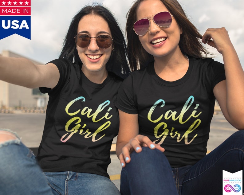 K&ouml;nnte beinhalten: Zwei Personen tragen schwarze T-Shirts mit dem Text "Cali Girl" in einem Regenbogen-Verlauf. Der Text ist in einer handschriftlichen Schriftart. Die Personen l&auml;cheln und tragen Sonnenbrillen.