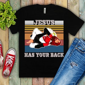 May include: Black t-shirt with a vintage-style graphic of a person in a jiu-jitsu hold. The graphic has a red, white, and blue color scheme and the text "JESUS HAS YOUR BACK".