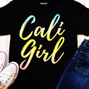 K&ouml;nnte beinhalten: Schwarzes T-Shirt mit einem Regenbogen-Verlaufs-Motiv, auf dem "Cali Girl" in einer stilisierten Schrift steht. Das Shirt wird mit einer blauen Jeans und wei&szlig;en Turnschuhen kombiniert.