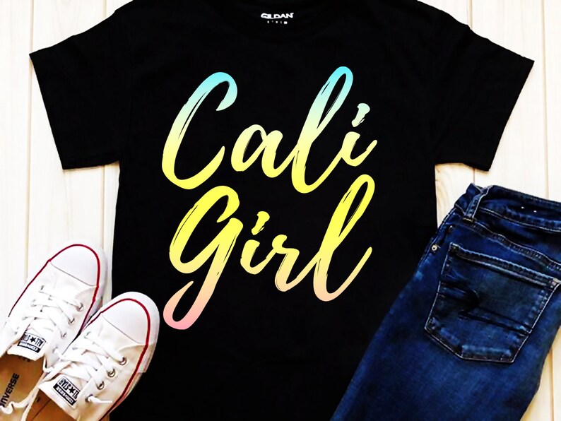 K&ouml;nnte beinhalten: Schwarzes T-Shirt mit einem Regenbogen-Verlaufs-Motiv, auf dem "Cali Girl" in einer stilisierten Schrift steht. Das Shirt wird mit einer blauen Jeans und wei&szlig;en Turnschuhen kombiniert.