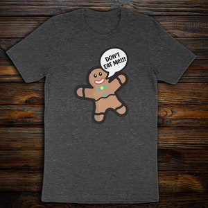 May include: A charcoal gray short-sleeve t-shirt featuring a brown gingerbread person graphic with a red smile, a blue bow tie, and a white speech bubble containing the black text "DON'T EAT ME!!!" printed on the front of the garment.
