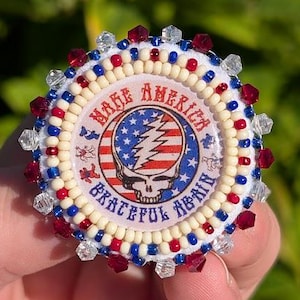 May include: A round, beaded pin with a central graphic featuring a skull, lightning bolt, and the words "Make America Grateful Again." The pin has a white, red, and blue color scheme, with clear and red beads around the edge.