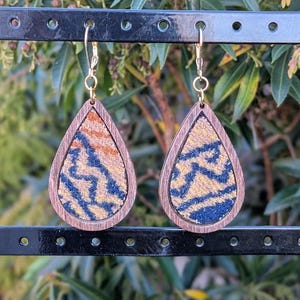 May include: Teardrop-shaped earrings with a wooden frame and a patterned fabric insert. The fabric features a design in shades of blue, tan, and orange. The earrings hang from gold-colored hooks, showcasing a unique and artistic design.