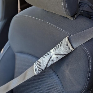 May include: A gray and black patterned seatbelt cover is shown on a car seatbelt. The cover is made of a soft material and features a geometric design. The car seat is black with white stitching.