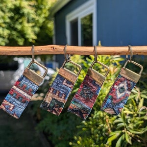 May include: Five colorful keychains with a woven fabric design and gold metal rings. The keychains are hanging from a wooden dowel.