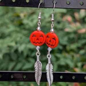 May include: A pair of dangle earrings featuring orange pumpkin-shaped charms with carved faces, silver feather charms, and silver crystal beads.