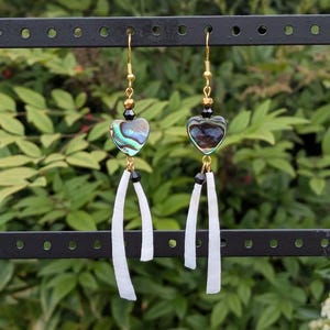 May include: A pair of dangle earrings with gold-colored hooks. Each earring features a heart-shaped iridescent abalone shell, black beads, and white, curved, rectangular pendants. The earrings are displayed against a green, leafy background.