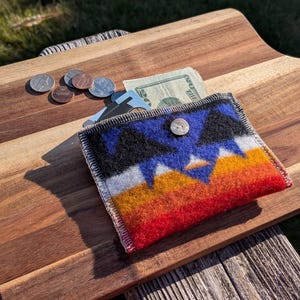 May include: A rectangular felt wallet featuring a geometric pattern in black, blue, white, yellow, and red. The wallet is secured with a single white button and contains a paper bill and a card, with several coins scattered nearby.