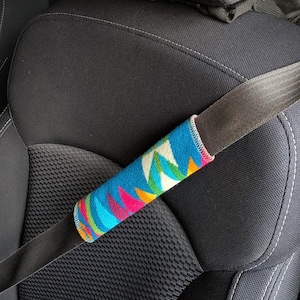 May include: A colorful seat belt cover with a geometric pattern in shades of blue, pink, green, and yellow. The cover is wrapped around a black seat belt, set against a black car seat.