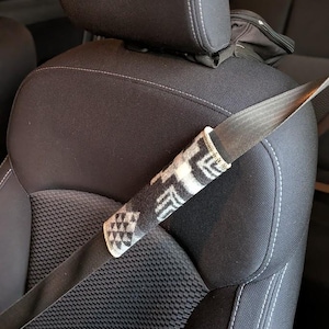 May include: A black and white patterned seat belt cover is wrapped around a car seat belt. The cover has a geometric design and is made of a soft, textured material. The car seat is black with white stitching.