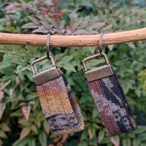May include: Two fabric keychains with metal hardware hang from a wooden dowel. The keychains feature woven patterns in shades of brown, orange, yellow, and black. The background is blurred green foliage.