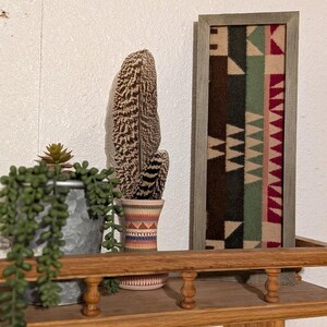 May include: A decorative shelf displays a framed textile art piece with a geometric pattern in brown, green, and red. A tall, patterned vase holds a feather, and a small succulent plant sits in a metal pot. The shelf is made of wood.