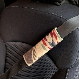 May include: A patterned seatbelt cover in beige, red, green, and black. The cover is cylindrical and wraps around a black seatbelt. The seatbelt is in a black car seat. The cover has a geometric design.