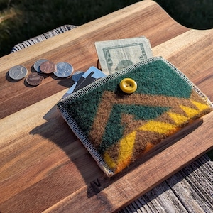 May include: A rectangular felt wallet featuring a geometric pattern in dark green, brown, and yellow. The wallet is secured with a single yellow button and contains a twenty-dollar bill and several cards, with loose coins scattered nearby.
