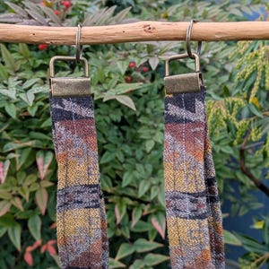May include: Two fabric earrings with a plaid pattern in shades of brown, yellow, and black hang from a wooden dowel. The earrings have a rectangular shape and are suspended by metal hooks.