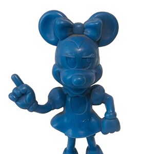 May include: A blue plastic figurine of a cartoon mouse wearing a dress and a bow. The mouse is pointing with its right hand and has a mischievous expression.