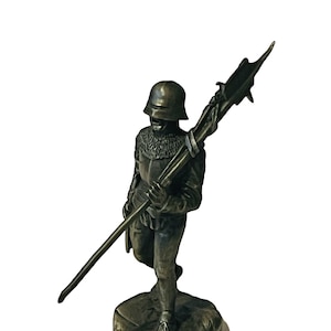 Puede incluir: Una figurita de bronce de un soldado con armadura y casco, sosteniendo una lanza con una punta de lanza. El soldado está de pie sobre una pequeña base.