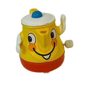 Puede incluir: Un juguete amarillo de cuerda con forma de tetera con una cara sonriente, detalles rojos y naranjas, y una tapa blanca con un pomo azul. El juguete tiene brazos blancos y una llave de cuerda en el lateral.