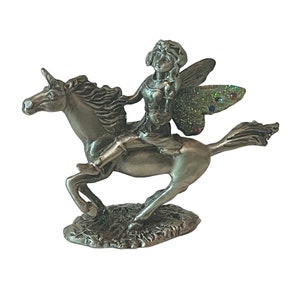 May include: A pewter figurine of a fairy riding a unicorn. The fairy has wings with glitter accents and is holding a flower. The unicorn has a horn and flowing mane.