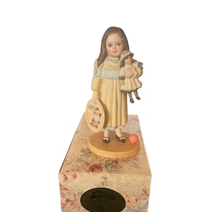 Könnte beinhalten: Eine Porzellanfigur eines jungen Mädchens in einem gelben Kleid, das eine Puppe hält. Sie trägt einen Strohhut und hat einen rosa Ball zu ihren Füßen. Die Figur steht auf einer Schachtel mit Blumenmuster.