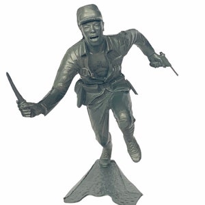 Peut inclure: Une figurine de soldat en plastique gris courant avec un couteau dans une main et un pistolet dans l'autre. Le soldat porte un chapeau et a une expression déterminée sur son visage. La figurine est debout sur une base en plastique gris.