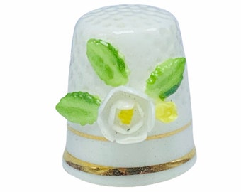 May include: White porcelain thimble with a gold trim and a white flower with yellow center and green leaves.