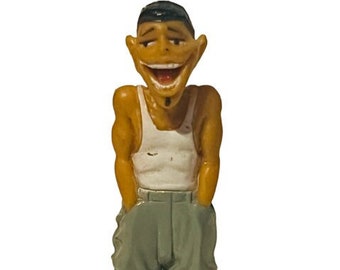 May include: A small figurine of a smiling man with brown hair, wearing a white vest top and grey trousers. He is standing with his hands in his pockets.