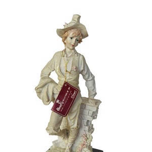 Puede incluir: Una figurita de porcelana blanca de un hombre con un sombrero y un bolso. La figurita está de pie sobre un muro de ladrillo y tiene una base con las palabras "Il Belcari" en ella. La figurita forma parte de la serie "Dear Collection of Art".