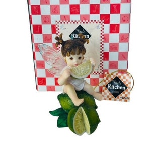 Könnte beinhalten: Eine Figur einer Fee mit Flügeln, die ein Limettenstück hält. Sie sitzt auf einem Bett aus Limetten. Die Figur ist Teil einer Kollektion namens "Little Kitchen Fairies".