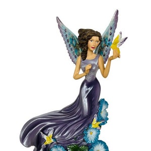 Peut inclure: Figurine de fée avec des ailes violettes et bleues, vêtue d'une robe violette fluide et tenant un papillon jaune. La base présente des fleurs de gloire du matin bleues et des feuilles vertes. La fée a les cheveux bruns.