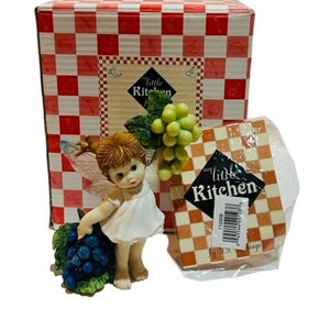 May include: A figurine of a fairy holding a bunch of green grapes. The fairy is wearing a white dress and has brown hair. The figurine is on a red and white checkered background with the text "my little kitchen" on a white tag.