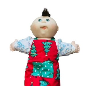 May include: A Cabbage Patch Kid doll wearing a red Christmas-themed apron with green trees and white polka dots. The doll has a brown mohawk, green eyes, and a pink nose.