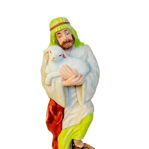 May include: A figurine of a person wearing a green and red robe holding a white lamb. The person has a green head covering and a beard.