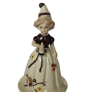 Könnte beinhalten: Eine Porzellanfigur eines Mädchens, das ein braunes Kleid und einen weißen Hut trägt. Sie hält ein kleines weißes Objekt in der Hand. Die Figur ist mit Blumen verziert und hat einen weißen Sockel.