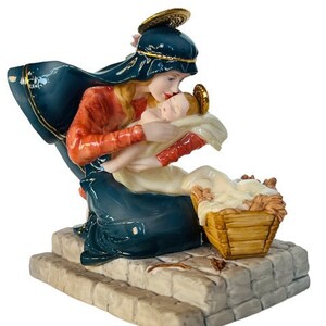 Op de afbeelding: Een keramieken kerststalbeeldje dat een vrouw in een blauwe mantel en een rode top toont die een baby vasthoudt. Het beeldje heeft gouden accenten en staat op een stenen voet. Een klein mandje staat naast de baby.