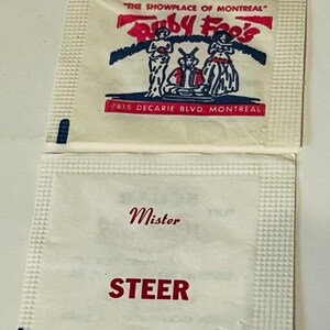 May include: Two white paper packets. The top packet has a red and blue logo that says "Ruby Foo's" and "The Showplace of Montreal" with the address "7815 Decarie Blvd. Montreal". The bottom packet has the words "Mister Steer" in red.